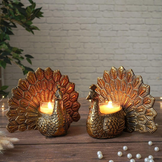 Two gold peacock-shaped candle holders on a wooden surface with a white brick wall background.