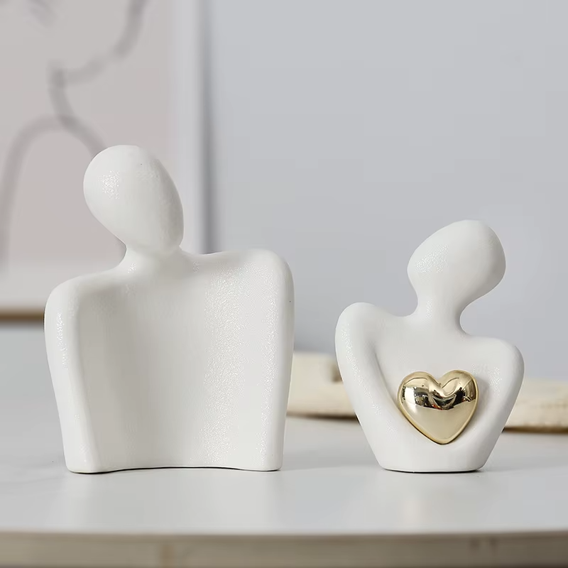 Two white ceramic figures, one with a gold heart detail, on a light surface.